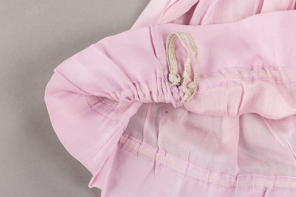 Hetten er rynket sammen i toppen og har rosett av silkebånd som pynt. Løpegang med strikk.