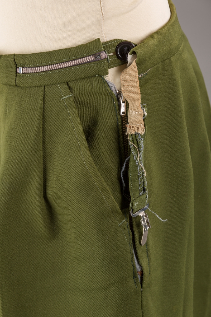 Grønn strekkbukse med stropper under bena. Avsmalnende ben. Glidelås i venstre side. Elastisk spennglidelås.