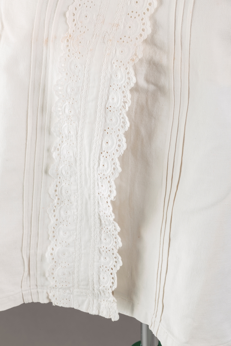 Form: Hoftelang trøye, Lange ermer, krage. Kragen og åpningen foran er kantet med  maskinprodusert blonde. Tre biser på hver side av åpn. foran. Lukkes med to løse knapper.