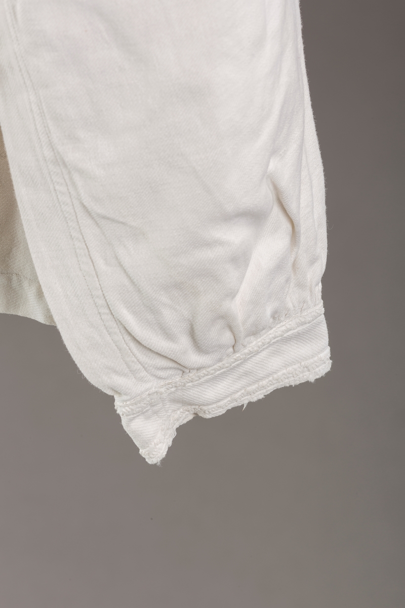 Form: Hoftelang trøye, Lange ermer, krage. Kragen og åpningen foran er kantet med  maskinprodusert blonde. Tre biser på hver side av åpn. foran. Lukkes med to løse knapper.