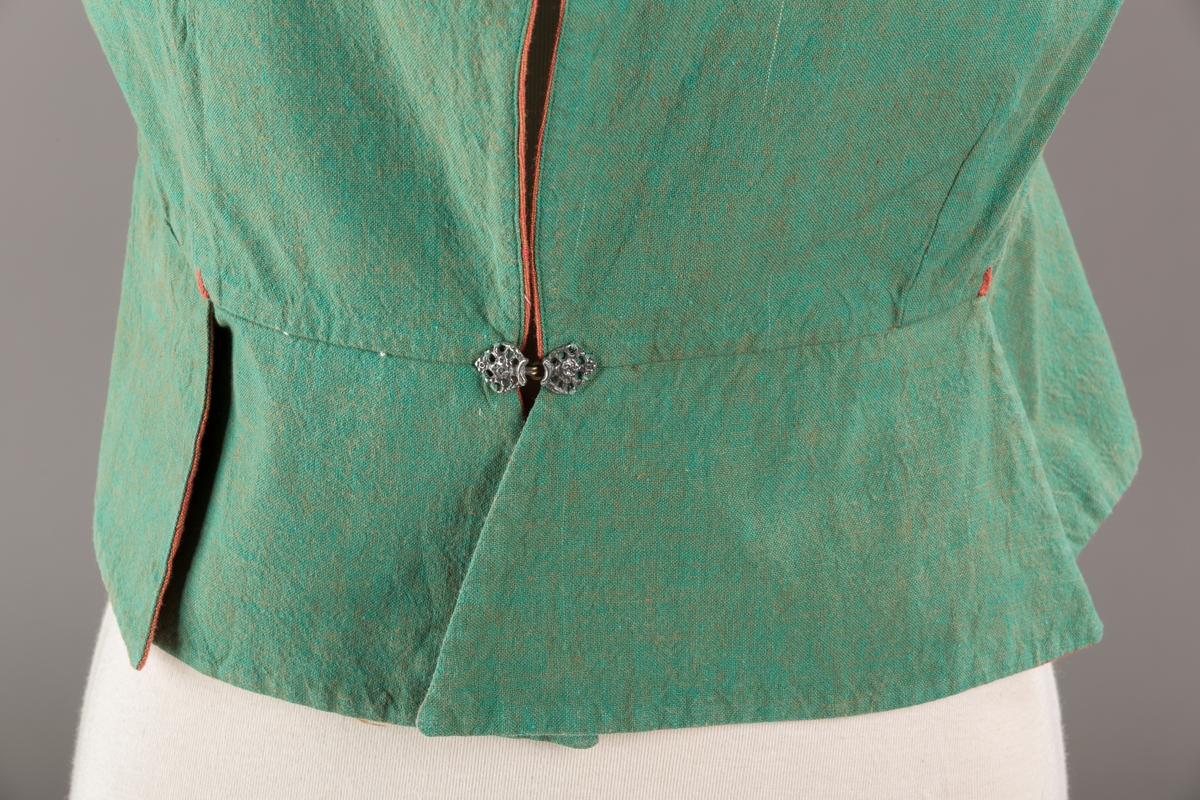 Grønt liv med rustrød kant og fór, seks skjøter. Håndsydd forsterkning i kontrastfarge øverst i skjøtesplittene. Lukkes med tinnhekte.