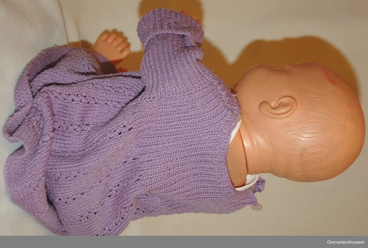 Plastik/gummi med bevegelige armer, hode og ben. Påkledd med babyklær: trøye og lilla strikket kjole.