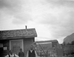 Kvinne, barn og mann foran gardsbruk. Motivet i nedkant av b