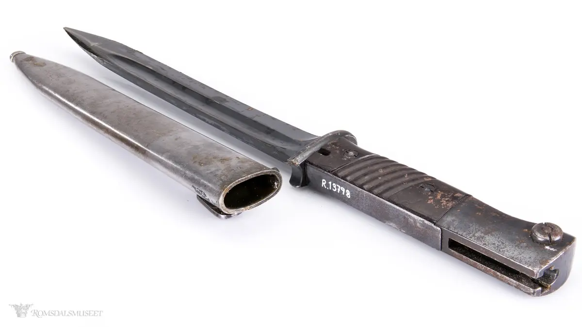 Metallhåndtak med lang pommel og geværfeste med fjæret knapp i bakkant. Serratert tregrep som er festet med 2 skruer. Langt spisst stålblad med egg og blodrand. Metallslire med kule enden og krok for festing til grunnutrustning/belte.