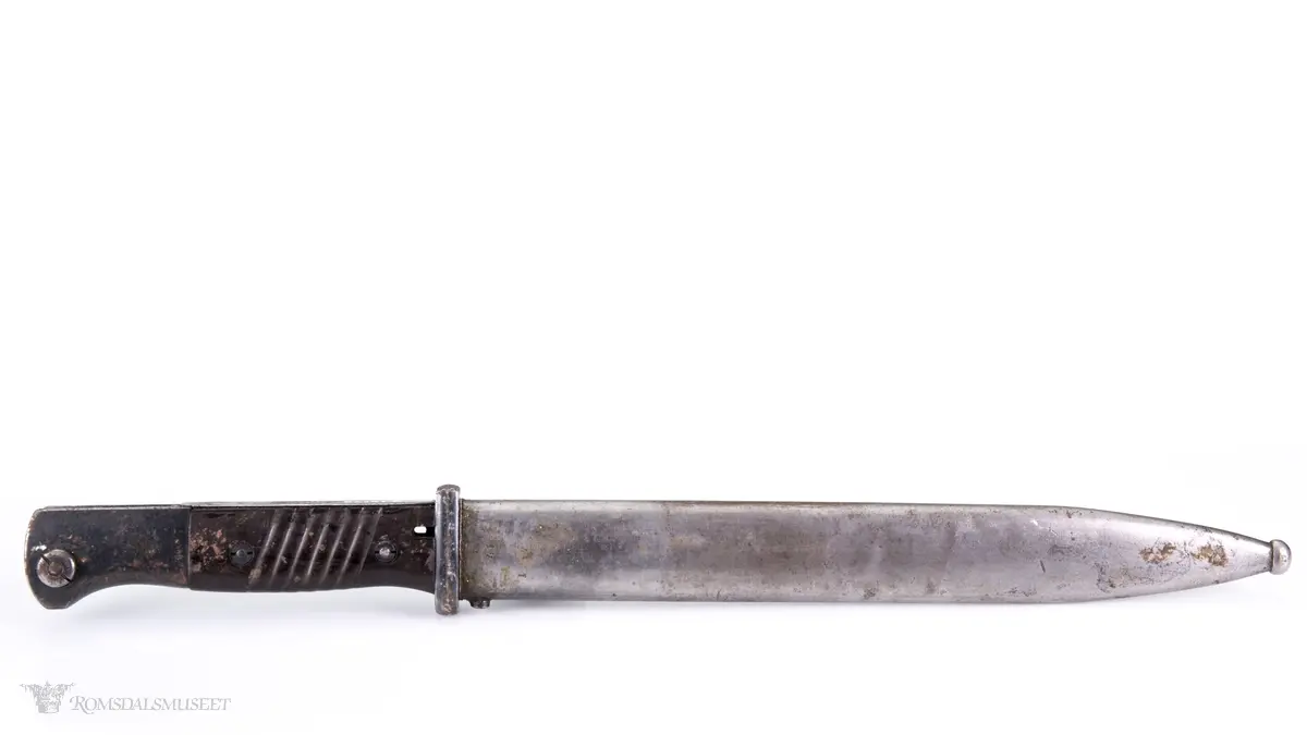 Metallhåndtak med lang pommel og geværfeste med fjæret knapp i bakkant. Serratert tregrep som er festet med 2 skruer. Langt spisst stålblad med egg og blodrand. Metallslire med kule enden og krok for festing til grunnutrustning/belte.