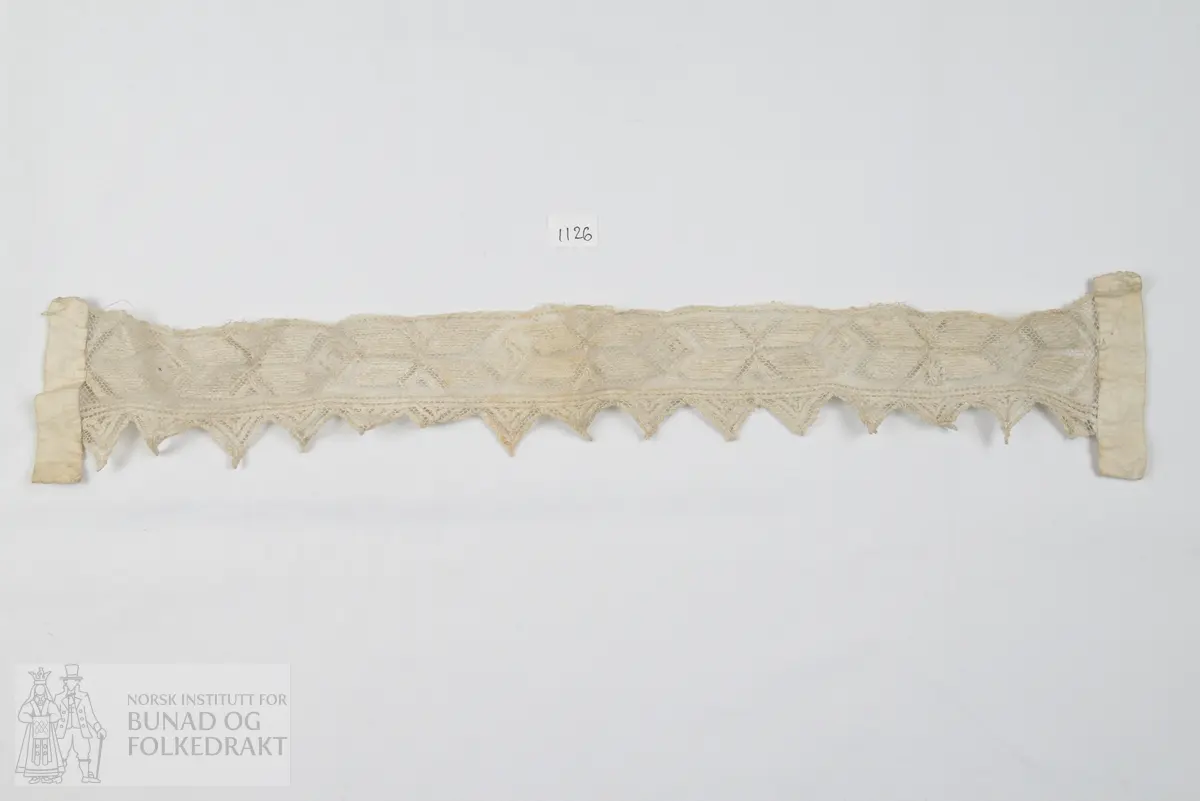 Ei lang remse av tyll forma med trekantsnippar etter eine langsida. Tyllstopping med grov laustvinna ujavn kvit lintråd - "gasisaum". I kvar ende er påsydd eit smalt dobbelbretta stoffstykkje av kvit linlerret.