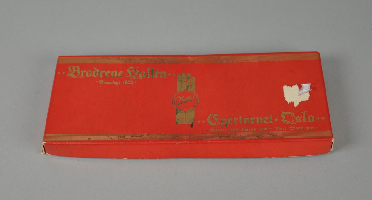 To lange brukte nylonstrømper i rød rektangulær eske med lokk og gulldekor.  Strømpene ligger i et hvitt silkepapir med samme tekst som på esken.