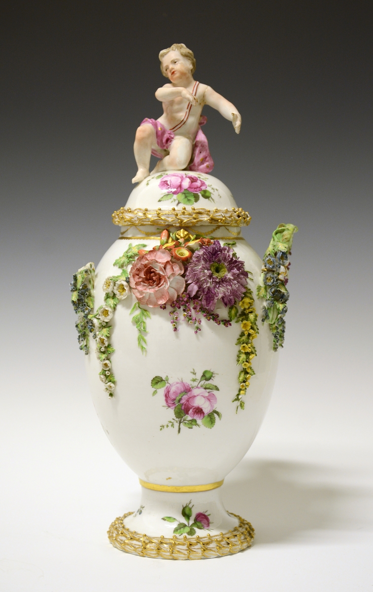 Potpourrikrukke av porselen med stett og lokk. Lokket er dekorert med håndmalt blomsterdekor og langs kanten en bord med blader i tre høyder dekorert med gull, over bladene gullfargede små kuler. På toppen av lokket en kjerub eller kupid som sitter på sitt ene kne. Han har på seg et rosa lendeklede og har holdt i et skjold eller en atributt. 
Urnens korpus er dekorert med håndmalte blomster, og modellerte blomster som sitter i klynger og strekker seg nedover korpus. De modellerte blomstene er uglasert og håndmalt. Urnen mangler en hanke. Den eksisterende hanken har modellerte blomster ved festene som strekker seg nedover i klynger.  Langs stettens kant samme bladbord som på lokket.