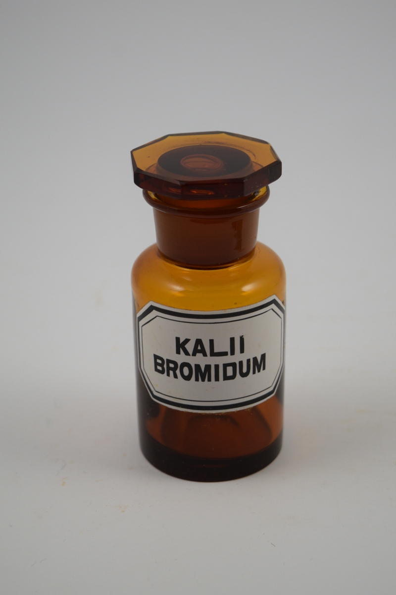 Brun glasskrukke med åttekantet glasspropp. Hvit etikett med sort skrift. Krukken ble brukt til oppbevaring av Kalii bromidum, som ble brukt som sovemiddel.