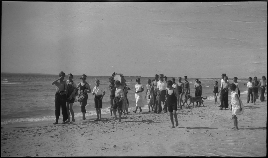 En stor vennegjeng i lystig lag på stranda på Sola. De har trommer og trekkspill og leker i vannet og på land med og uten ball. Sola strandhotell ligger i bakgrunnen. Tor Johannessen og Harald Bergsaker er på bilde nr. 14 og 18.