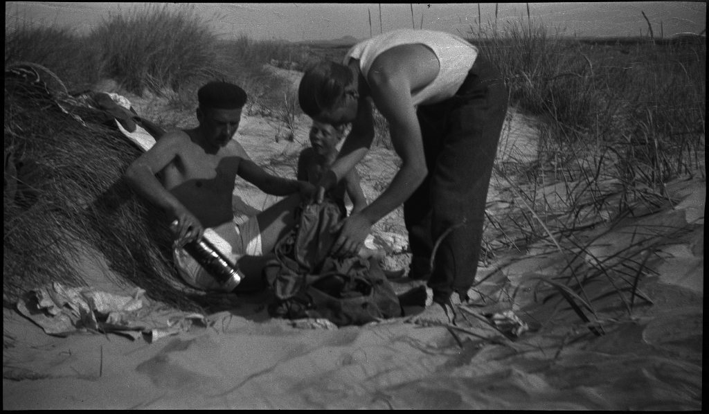 Unge menn og gutter soler seg i sanddynene på Solasanden. To gutter leker med en kikkert. Lindtner har svart alpelue på.