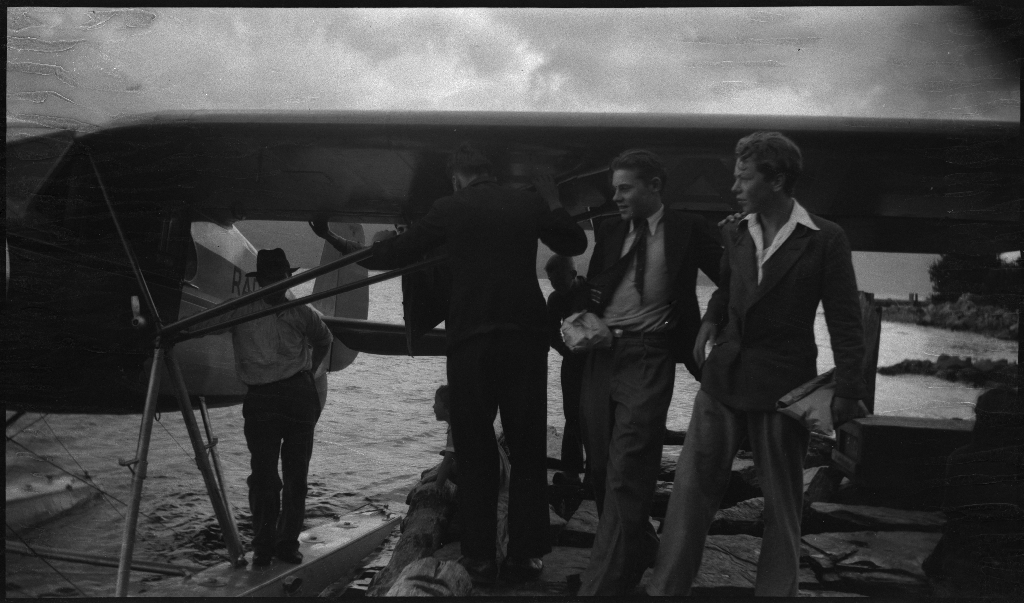 Alf Olaf Ådnøy og Egil Ellingsen står foran et lite sjøfly med påskriften "Radionette LN-EAF". Mange menn og barn studerer flyet, og tre kvinner står ved et hus og ser på.