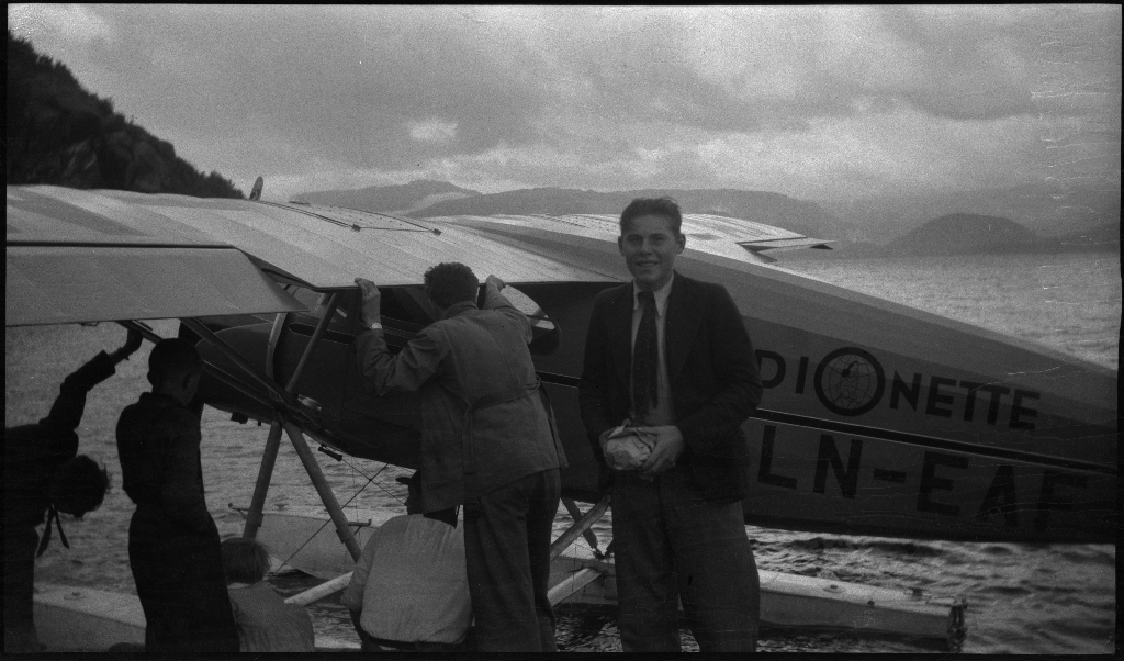 Alf Olaf Ådnøy og Egil Ellingsen står foran et lite sjøfly med påskriften "Radionette LN-EAF". Mange menn og barn studerer flyet, og tre kvinner står ved et hus og ser på.