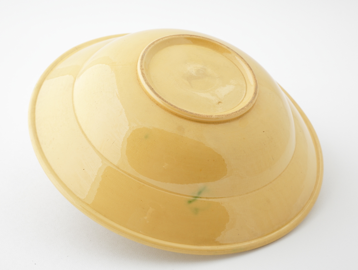 Skål i glaserad keramik.
Rund skål med honungsgul glasyr, försedd med invändig dekorrand (kedja) i brunt och två gröna ränder i rinningsglasyr. I botten en cirkelrund grön rand.