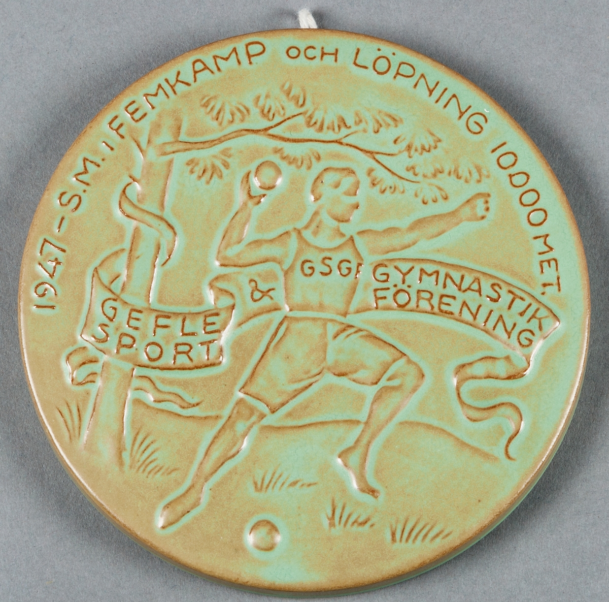 Väggmedaljong till minne av SM i femkamp och löpning 10 000 meter 1947, arrangerat av Gefle Sport & Gymnastikförening GSGF. Tillverkad av Bo Fajans AB i Gävle.