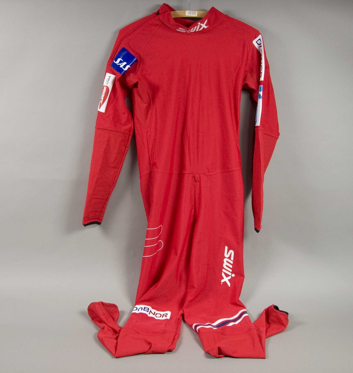 Rød langrennsdress med hvite og blåe striper. På dressen er det flere laminerte logoer for forskjellige sponsorer.