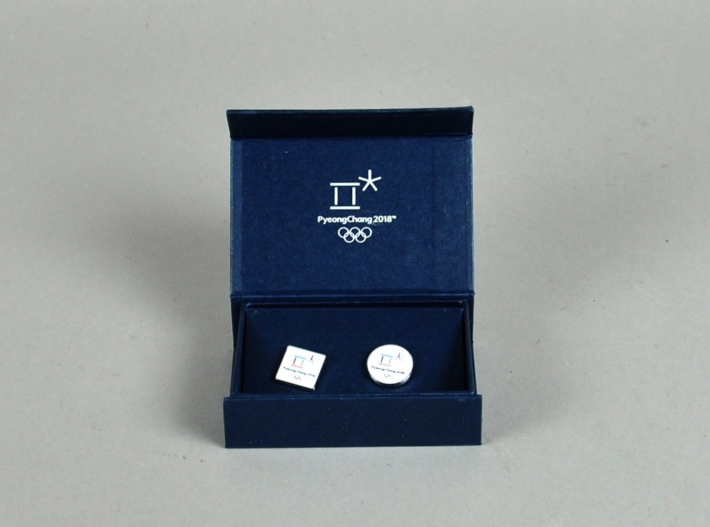 To mansjettknapper, en rundt og en kvadratisk, begge med logo for de olympiske vinterlekene i PyeongChang 2018 på hvit bakgrunn. Mansjettknappene ligger i et blått etui med logo i sølv.