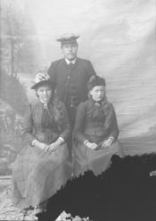 Mann står bak to kvinner som sitter på stol. Bilde er svart 