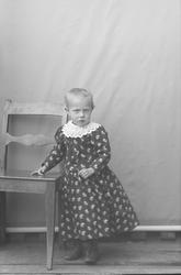 Portret. Lita jente står ved stol. Ca. to-tre år.