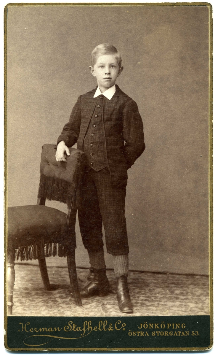Kabinettsfotografi: okänd pojke i 10-årsåldern stående med handen på stolsryggen vid ett bord. Klädd i svagt rutig jacka, väst och byxor, som slutar strax nedanför knät.