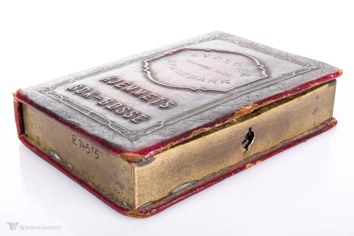 En sparebøsse i messing, formet som en liten bok, kledd i skinnimmitasjon.