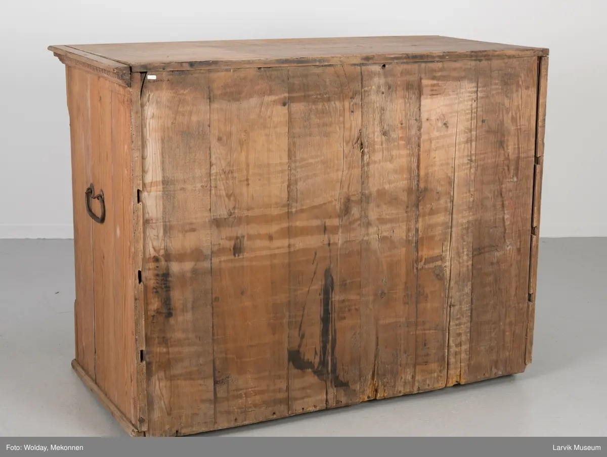 Kommoden har 4 skuffer. 2 beslag for å dra ut, 1 beslag for nøkkelhull, messing. Utskåret dekor under øvre pyntelist "tanngard" rundt på 3 kanter. Øvre skuff utskåret med bølgebånd med rett linje som bryter bølgene. Utskjæring kun i front. To av kommodens hjørner har utskåret søyle. Håndtak på kortsidene for å bære i.