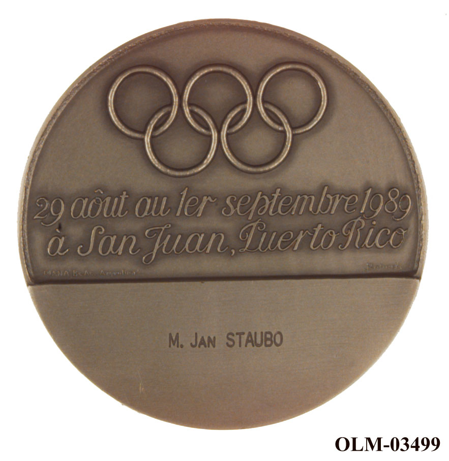 Medalje med et skjoldlignende motiv på den ene siden og de olympiske ringer og tekst på den andre siden. Medaljen ligger i en treeske.
