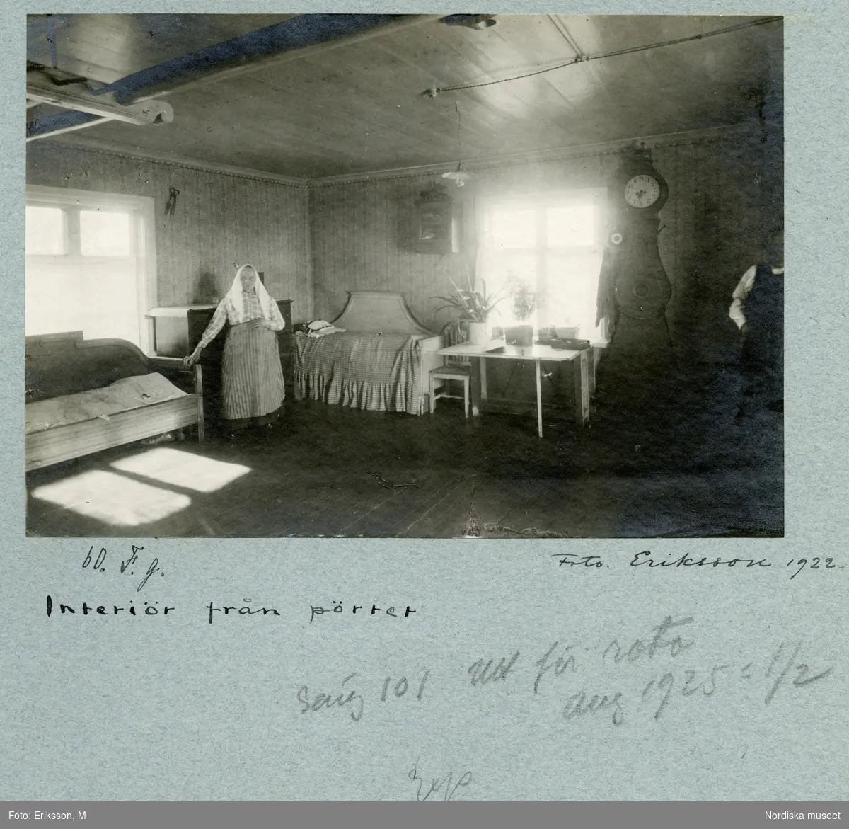 Interiör från pörtet. En kvinna står och håller sig i en av de två sofforna som står längs väggarna. Vid fönstret ett skrivbord och ett golvur.