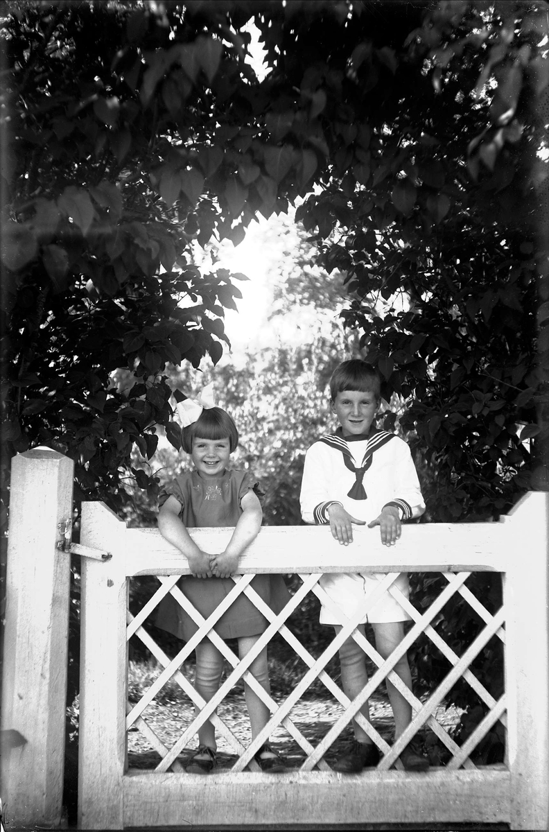 Barnen Renström. Georg Renströms barn Folke och Margot i sommarkläder bakom trädgårdsstaket.