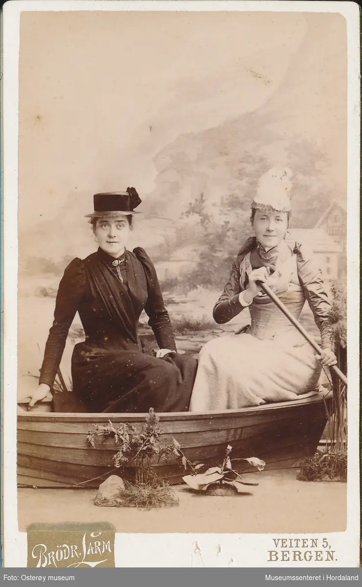 atelierfotografi av to unge kvinner i ein robåt framfor malt landskap. Kvinna til venstre i mørk kjole og hatt, den andre i ljos kjole med høg kvit hatt