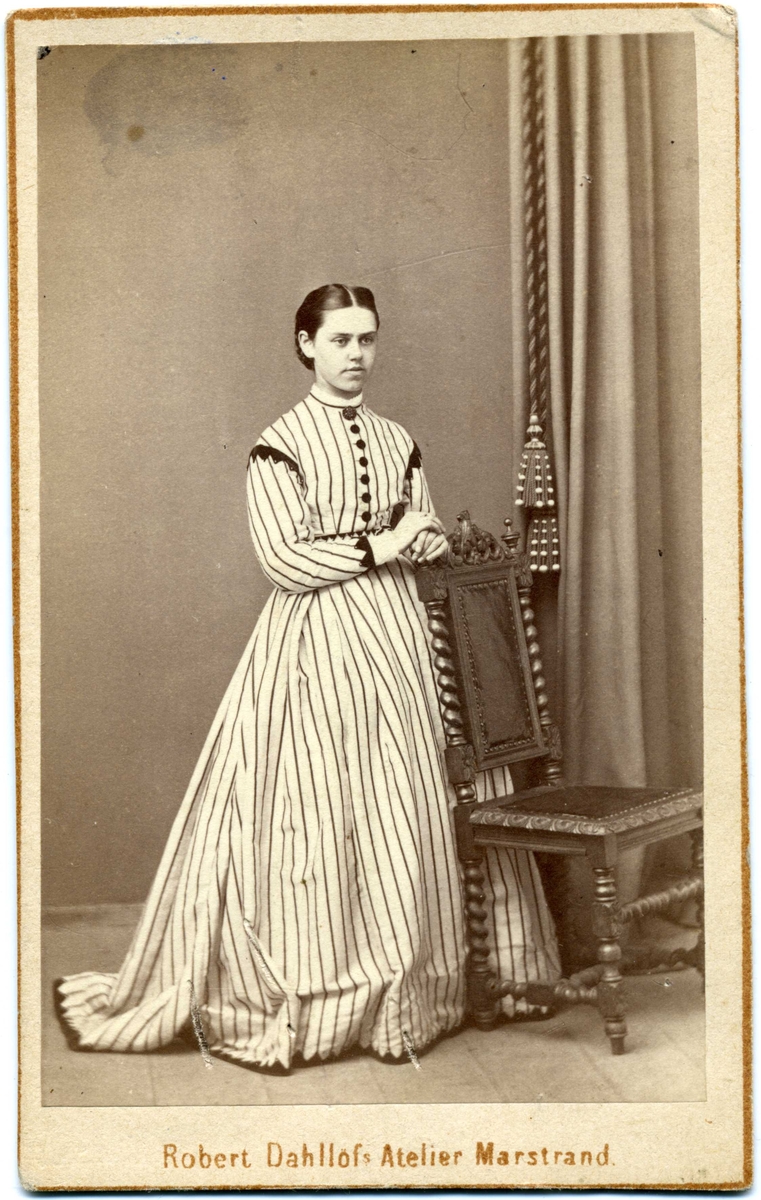 Kabinettsfotografi: okänd ung kvinna i randig klänning med markanta mörka knappar vid bröstet. Hon står, vilande med händerna mot en stolsrygg.