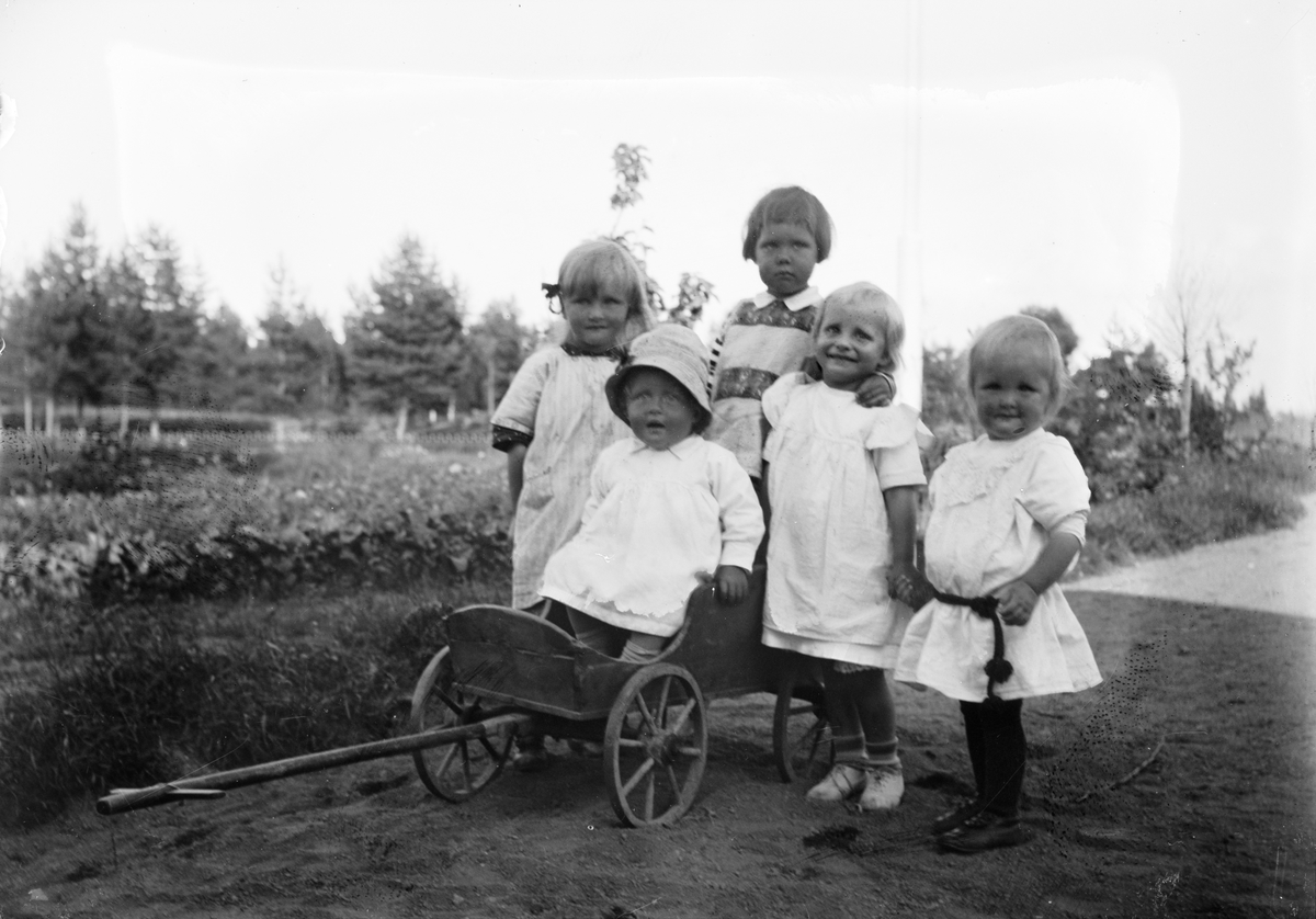 Marit Liljefors med fyra andra små flickor i trädgård, en sitter i en kärra. Sannolikt i Sverige