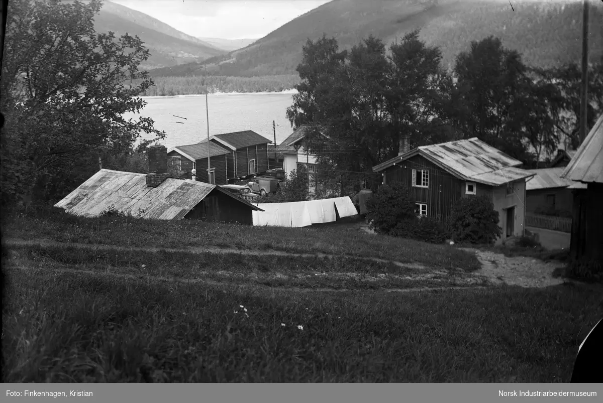 Bolighus ved Sigurdsrud med utsikt til Tinnsjøen. Tøy er hengt ut til tørk. Biler stående på gårdsplassen mellom hus og uthus.