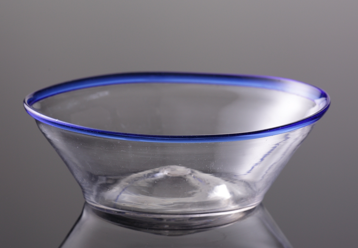 Filbunke av glas, med omvikt blå kant.
Det blå glaset "spinner" man på innan mynningskanten drivs ut till önskad form.