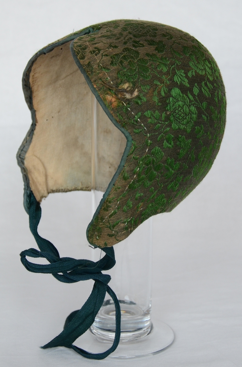 Bindmössa, så kallad finka, klädd med mönstervävt bomullstyg i brunt med blommor i grönt silke. Knytband av grön sidenrips. Fodrad med linnelärft.