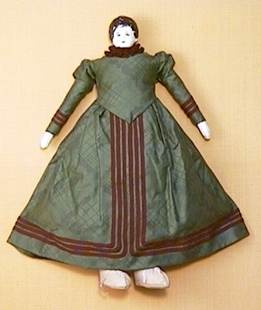 Docka med porslinshuvud. Sydd omkring 1970 med kläder i 1890-talets stil. Grön klänning av bomullsatin, fyra underkjolar, mamelucker. Snörskor av ljust skinn.