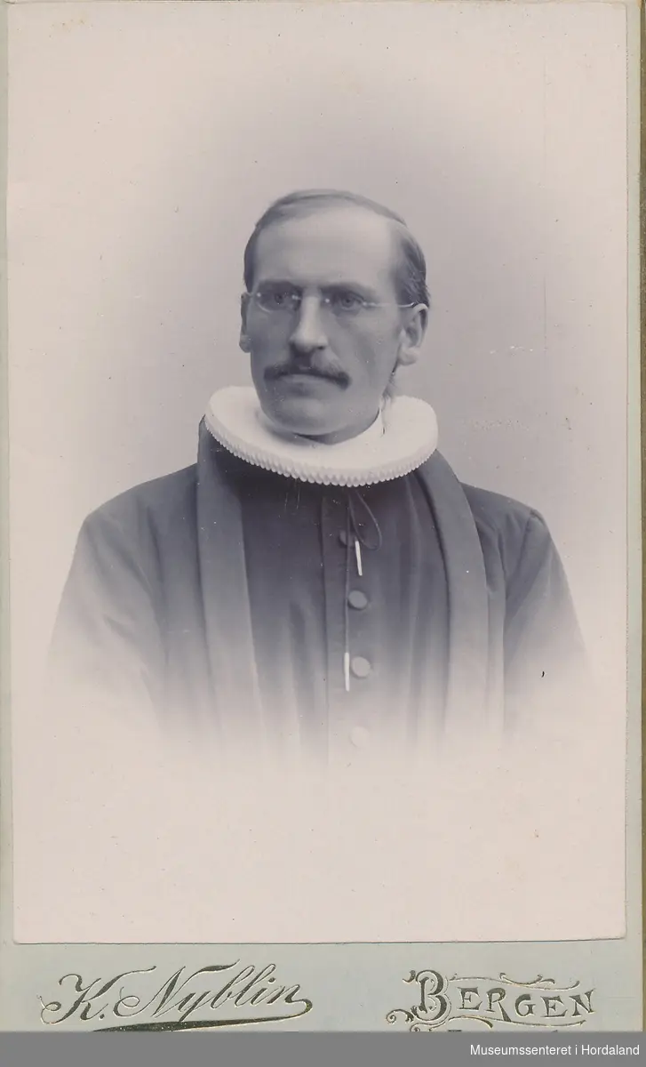 Halvfigurfotografi av mann med briller, tynt hår og bart, kledd i embedsdrakt med pipekrage.