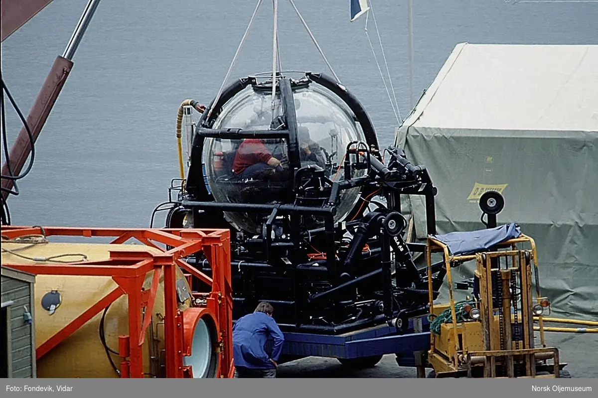 To menn sitter i miniubåten Check Mate, utviklet av NUI/NUTEC.  Ubåten står på kaikanten ved NUI's base, og er festet til en kran.