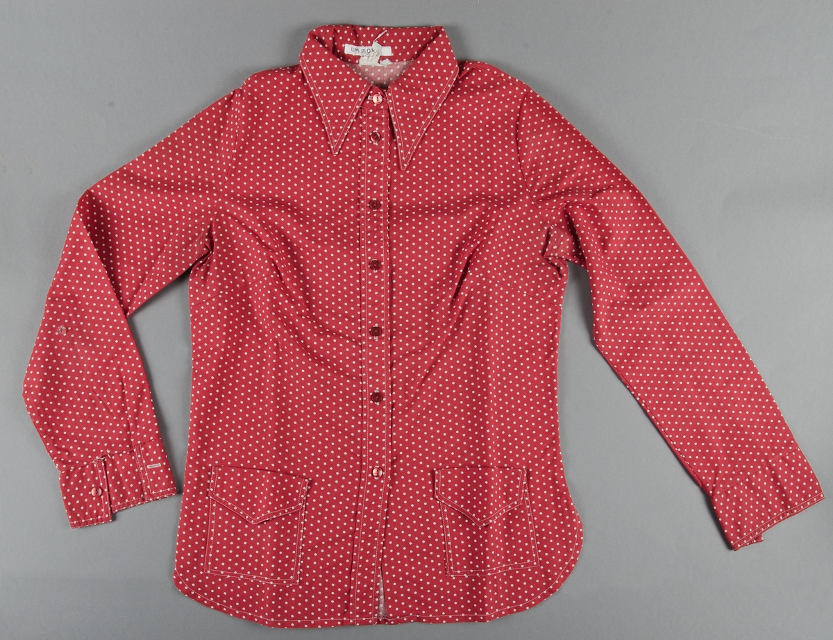 Blus, s.k. utanpåskjorta, röd- och vitprickig av bomull. Påsydda fickor med ficklock fram. Röda plastknappar. Total längd 70 cm. Ärmlängd 59 cm. Axelbredd 36 cm.