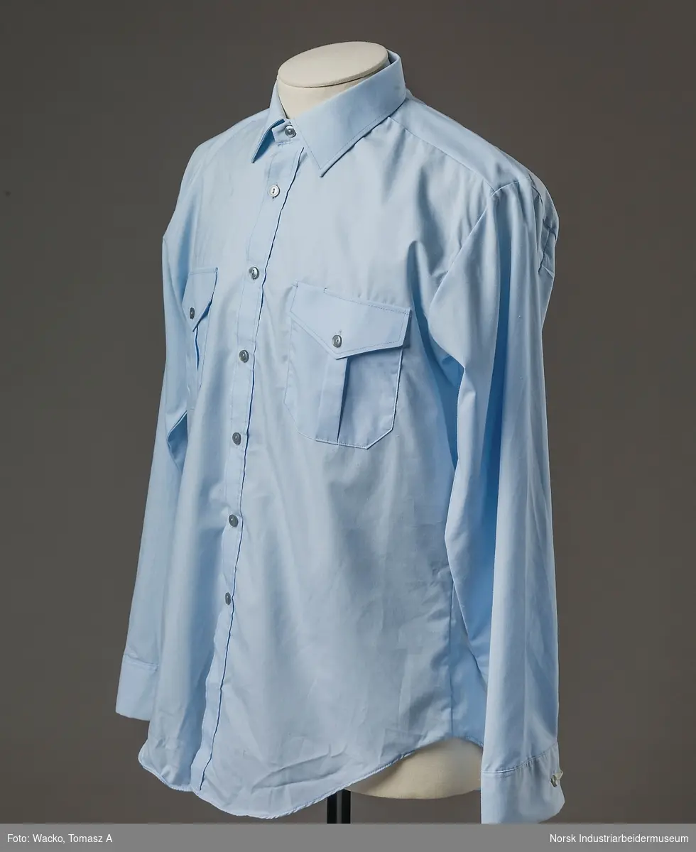Lys blå, langermet uniformsskjorte i tekstil av 65% polyester og 35% bomull. Nedfelt krage. Lukking i front med 7 grå knapper. Brystlommer med klaff som lukkes med en grå knapp. Mansjett har et knappehull og lukkes med grå knapp.