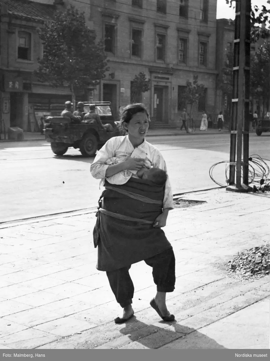Koreakriget. Koreansk kvinna ammar sitt spädbarn stående på en gata. I bakgrunden amerikanska soldater i en jeep.