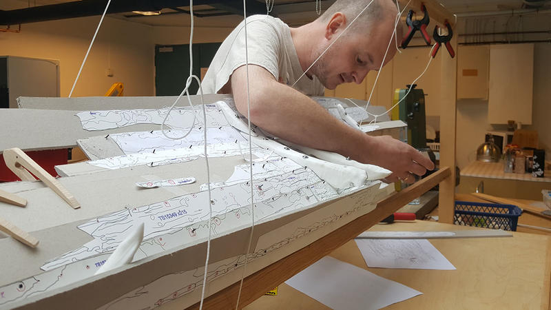 Arkeolog bøyer seg over og arbeider på innsiden av en uferdig båtmodell i plast og papp. Modellen er støttet opp i en treramme ved hjelp av klemmer og klyper i tre og plast, samt snører på flere steder.