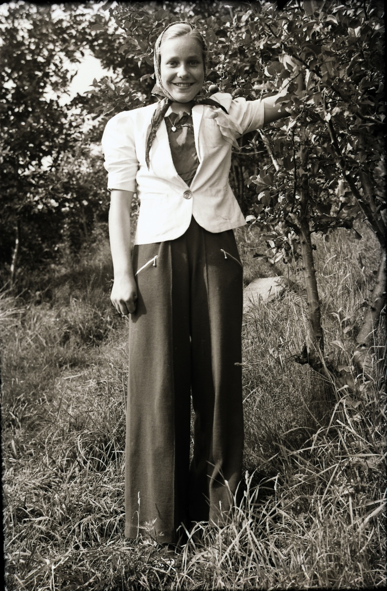 Anna-Lisa i Hugalyckan står vid ett fruktträd, poserande i modernt vida byxor, vit kort kavaj med puffärmar, halsband med hjärtberlock och en sjalett om huvudet.
(Se även bildnr EA0297)