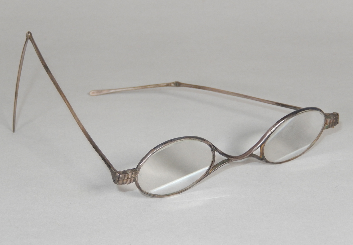 Glasögon med ovala glas och metallbåge. Skalmarna består av två delar som kan fällas ut och böjas. Glasögonfodral, avlångt, av trä med rundade hörn. En del uppfällbar, metallskodd, för att kunna ta ut glasögonen.