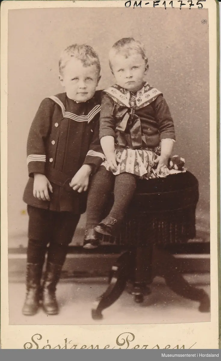 fotografi av to små born, gut og jente, med ljost hår, han i matrosdrakt og ho med matrosliknande kjole med mønster, mørke strømper og svarte sko
