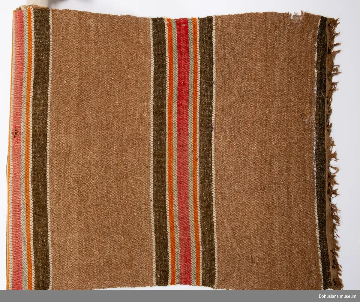 Beige-brun. Tvärrandig i rött, ljusblått, orange, vitt och grått.
Denna matta är en av fyra stycken som låg i en fyrkant i salen hos givarens mormors mor på Orust från ca 1890 och framåt tiden.
Se bilaga i Bilagepärmen.