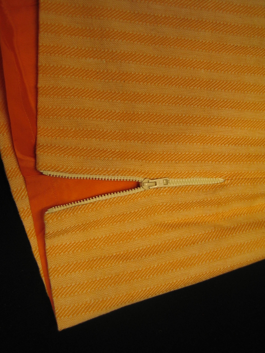 Orangegul sidenfodrad rak kjol i bomull. Tyget är vävt i kypert och tuskaft i 10 mm breda ränder. Kjolen har två sprängveck bak och dragkedja i vänster sida och en 25 mm bred linning med knapp och knapphål. Kjolen är försedd med ett sprund med dragkedja i vänster sida.