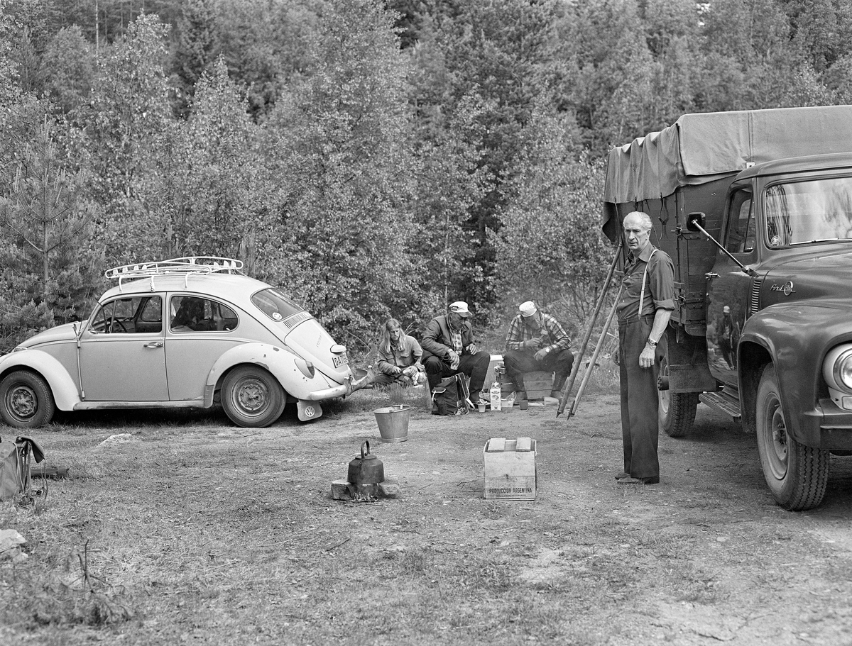 Spisepause for tømmerfløterne ved Kvernfallet i elva Søndre Osa i Åmot i Hedmark i 1981. Lastebilen til høyre ble brukt til å transportere utstyr og mat. Paul Solberg, som står til høyre på dette bildet, kjørte bilen. Han hadde også ansvaret for kaffekoking og liknende. Her hadde han tent et lite bål mellom to steiner og kokt kaffe på en sotsvart kjele. Til venstre, bak kaffebålet, ser vi Kenth Mokkelbost fra Flisa i Åsnes, som akkurat da dette fotografiet ble tatt satt og skar ei skive fra et brød han hadde lagt på låret. Ved siden av sitter Arne Norsted og Arnstein, Norsted, far og sønn, fra Osen. på hver sin trekasse. Også de spiste da bildet ble tatt. Lastebilen var en amerikansk Ford, antakelig en modell fra perioden 1953-1955.  Inntil bilen sto det to fløterhaker. Folkevognbobla til venstre i bildet er sannsynligvis fra slutten av 1960-åra.