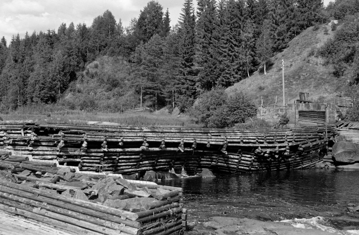 Syversætredammen i elva Flisa i Åsnes i Solør, fotografert fra nordvestre elvebredd i 1955.  Bildet ble tatt i en tørkeperiode midt på sommeren, da vannføringa i elva var minimal, slik at damkonstruksjonen var godt synlig, uten overløpende vann.  Dermed ble forfallet og råteskadene på tømmerkonstruksjonen, som åpenbart var moden for fornyelse, lett synlige.  Syversætredammen var en terskeldam som var bygd øverst i fossen, åpenbart for å skape et vannreservoar for det nedenforliggende kvernbruket, muligens også for å heve vannspeilet og lette tømmerfløtinga i det ovenforliggende elveløpet.  Dammen skapte et fall på cirka to meter.  Også nedenfor denne konstruksjonen rant vannet, hvert fall i flomperioder, kvitskummende med et visst fall.  Da dette fotografiet ble tatt var vannføringa svært liten og vannet i elveløpet var forholdsvis stillestående.  I forgrunnen til venstre ser vi enden av ei steinfylt tømmerkiste, antakelig en såkalt skådam som fungerte som avgrensning for et tømmerløp ved nordvestrere elvebredd.  På motsatt side av elva ser vi en damkonstruksjon av stein, antakelig for inntaket for driftsvann til kverna som lå på den sørøstre sida av elva.  SJF.1989-02316 viser den samme dammen, sett fra omtrent samme opptaksposisjon, mens den ennå var i god stand og på et tidspunkt da det var mye vann i Flisa.