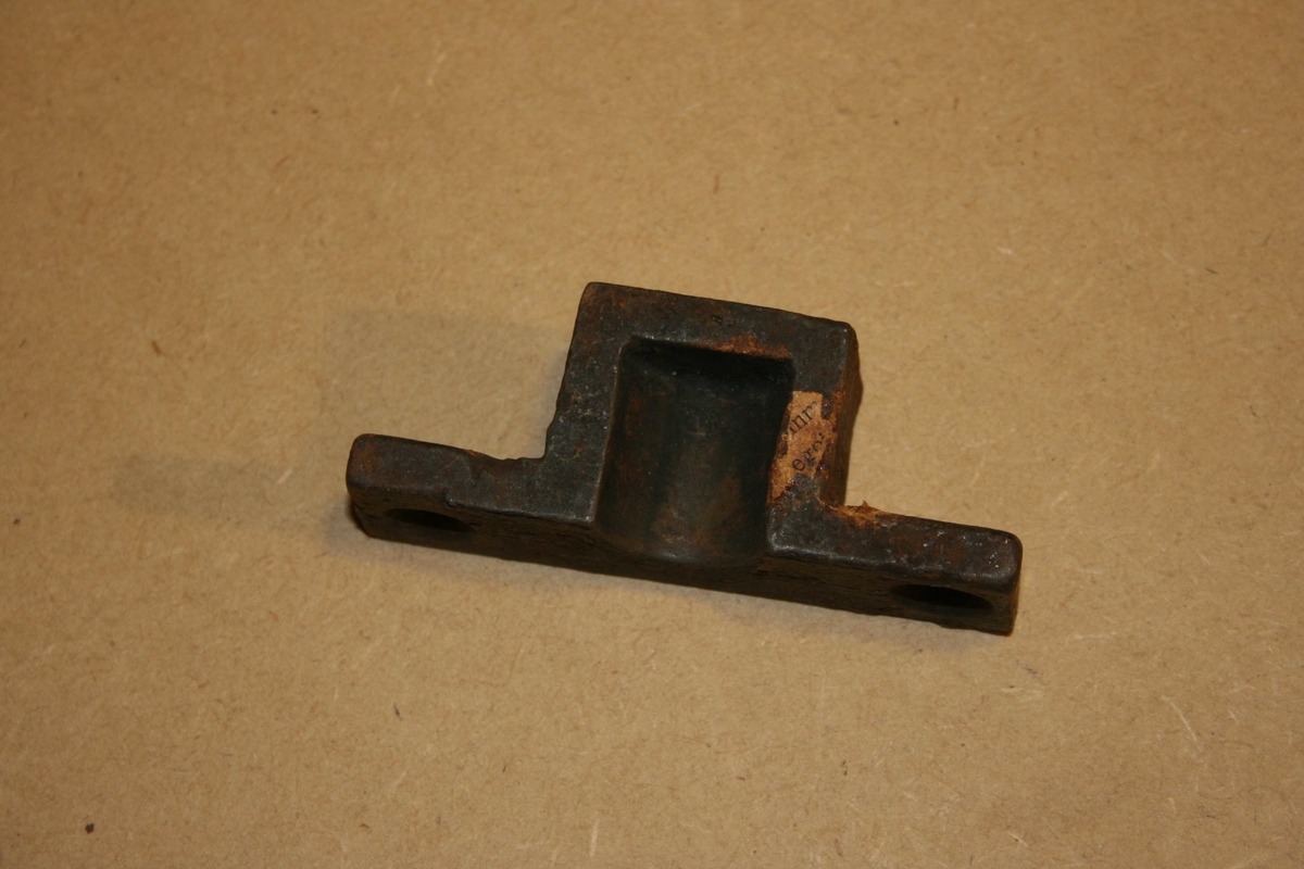Rektangulært jernstykke med firkanta forhøgning midt på.Halvsylinderforma utsparing på sida . 2 runde hol. Rusta.